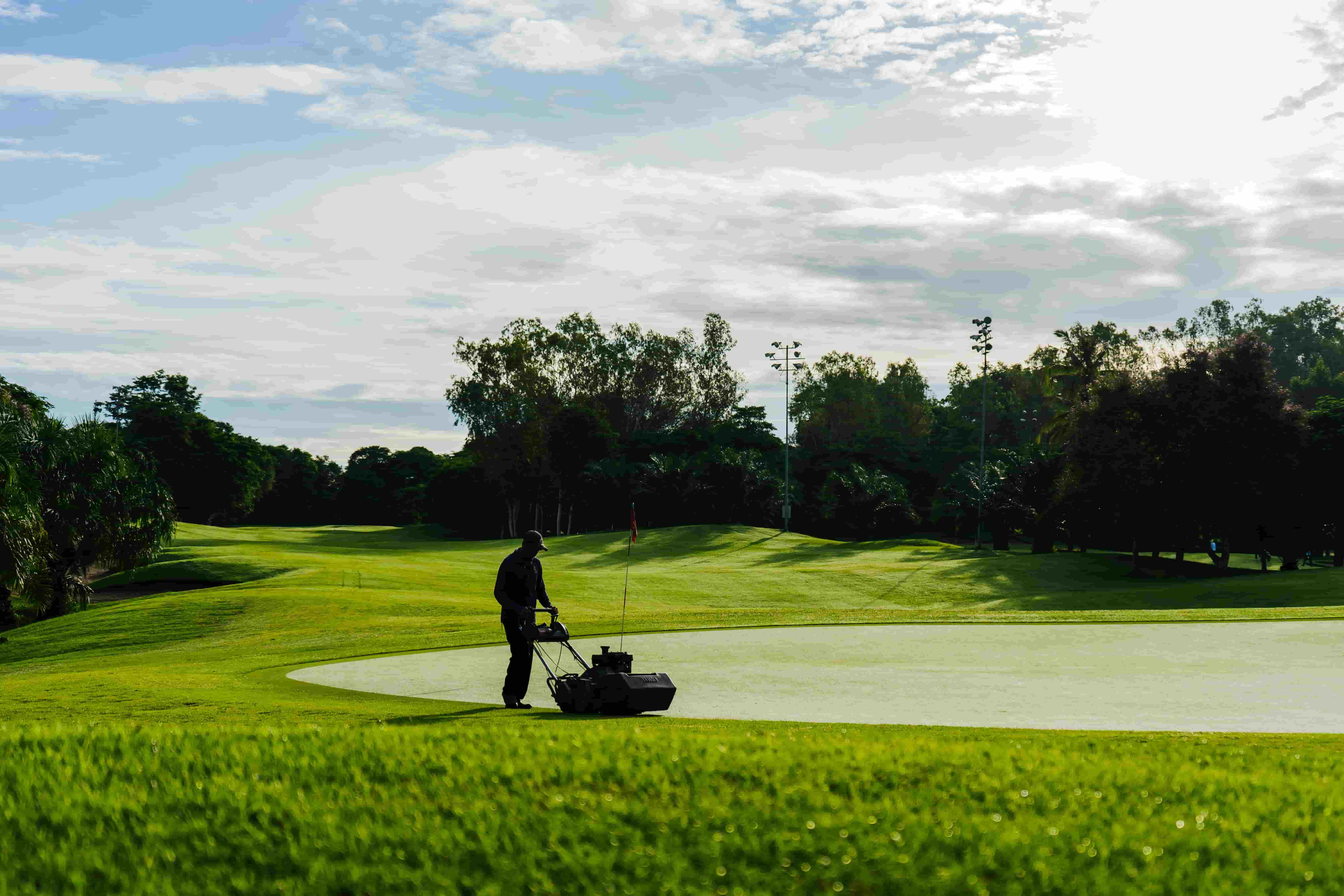 Groundskeeper pushing a reel mower across a manicured green on a sunny morning