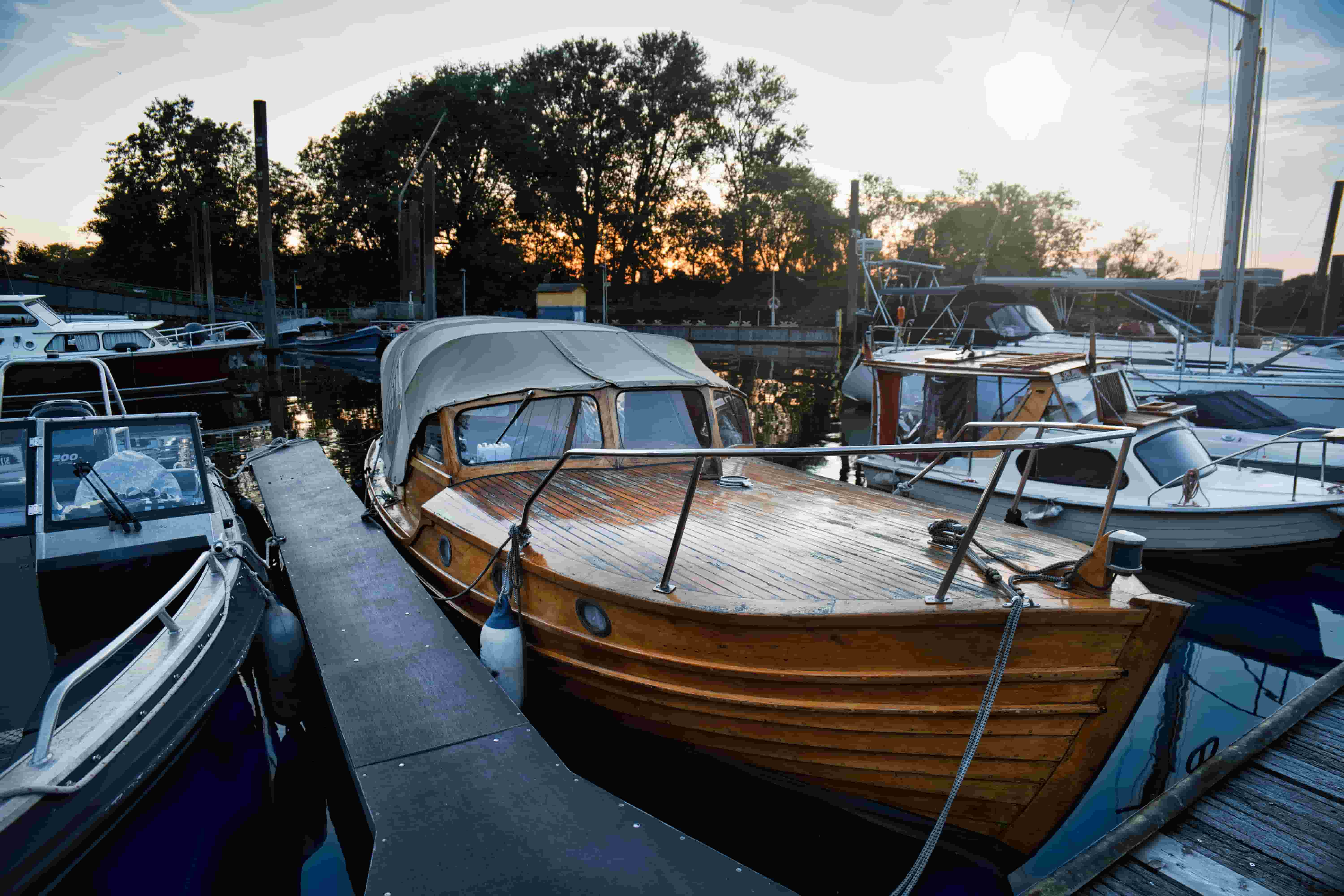 Varnished wooden runabout under a canvas cover tied up at a marina dock at sunset, with powerboats and sailboats in the slips around it